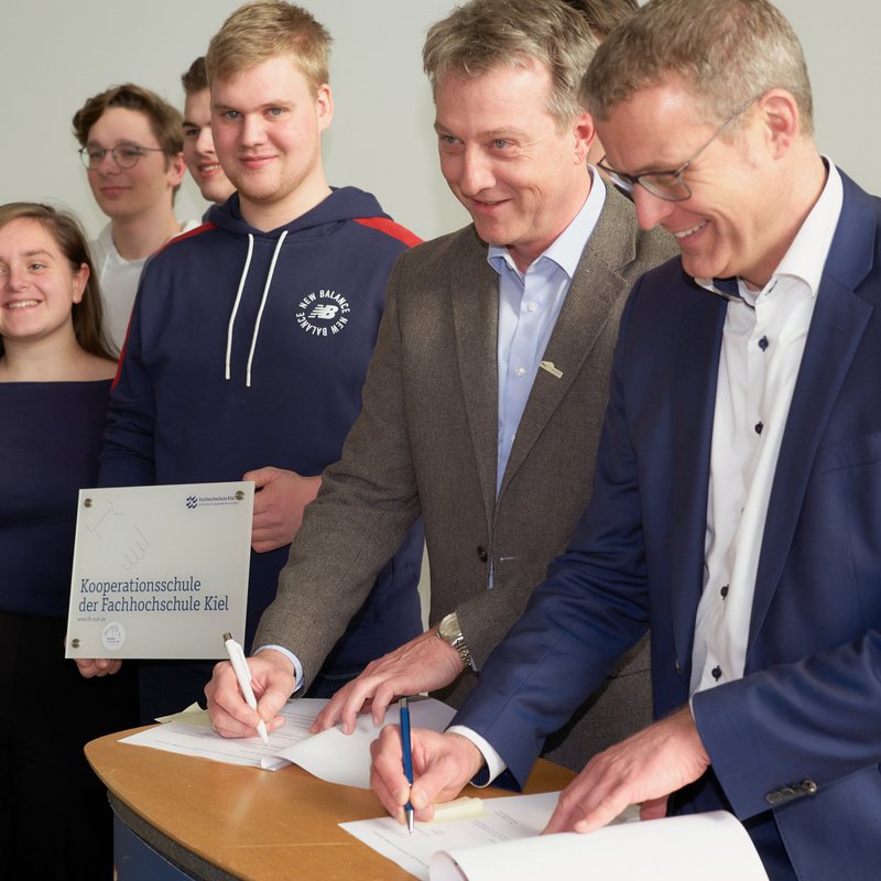 Prof. Christensen, Präsident der FH Kiel und Jörg Schelzig (Schulleiter RBZ Technik Kiel) unterzeichnen einen Kooperationsvertrag. Im Hintergrund sind Schüler*innen zu sehen.