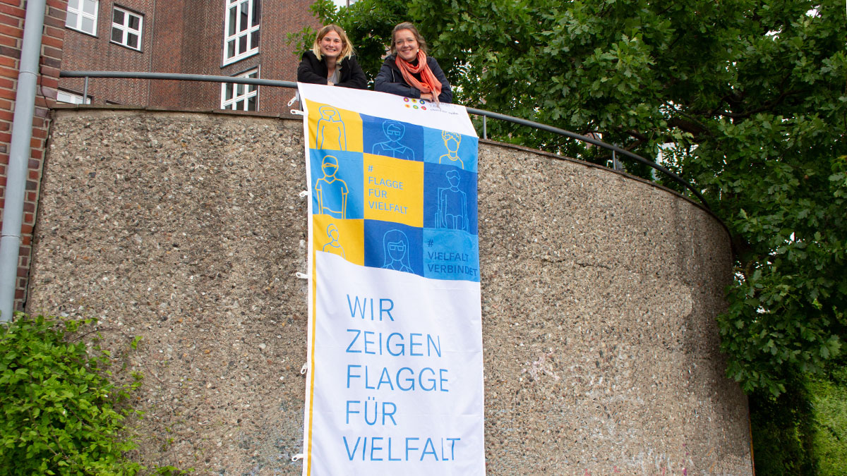 Zwei Frauen schauen lächelnd in die Kamera, vor ihnen hängt eine Flagge mit der Aufschrift: "Wir zeigen Flagge für Vielfalt"