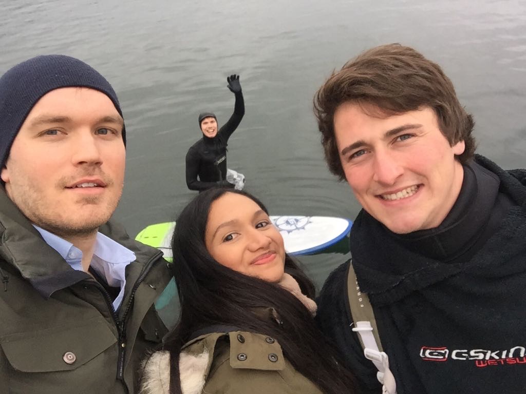 Zwei Männer und eine Frau machen ein Selfie am Ufer, auf dem Wasser winkt ein Mann in die Kamera.