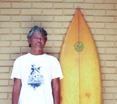 Ein Mann in weißem T-Shirt, steht neben seinem gelben Surfbrett.