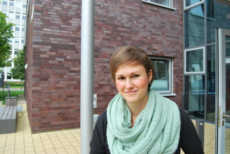 Ein Frau mit kurzen Haaren und türkisem Schal, schaut vor einem Gebäude der FH Kiel in die Kamera.