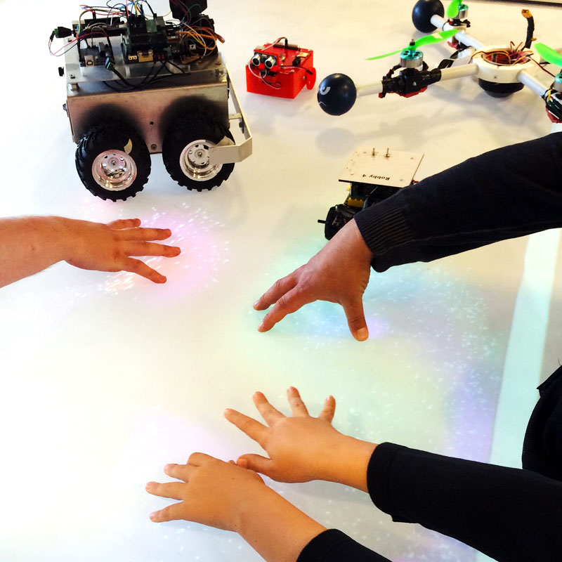 Kinder berühren den Multitouch-Tisch, auf dem kleine Roboter stehen
