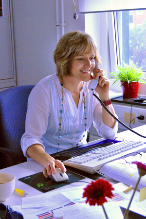 Eine Frau in weißer Bluse sitzt beschäftigt, aber glücklich, an ihrem Arbeitsplatz. In der einen Hand hält sie das Telefon, in der anderen die Maus.