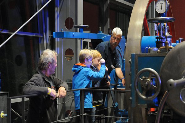 Zwei Jungen dürfen unter Aufsicht zwei erfahrener Männer einen Hebel an einer großen Maschine bedienen.