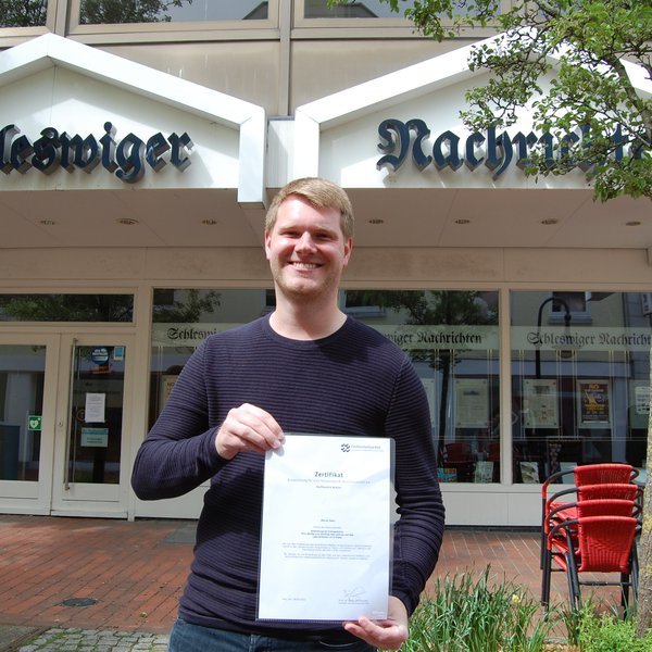 Marcel Nass steht mit seiner Auszeichnungs-Urkunde vor dem Gebäude der Schleswiger Nachrichten