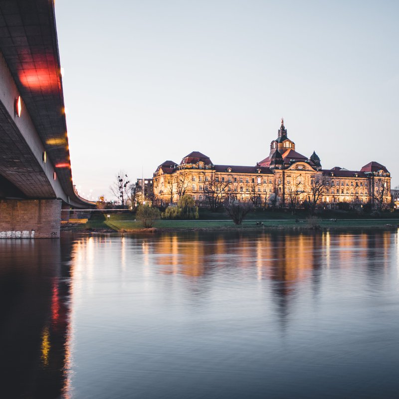 Auf der linken Bildseite ersteckt sich die Unterseite einer Betonbrücke, am gegenüberliegenden Ufer des Flusses, in diesem Fall der Elbe, ist die Sächsische Staatskanzlei zu sehen.     