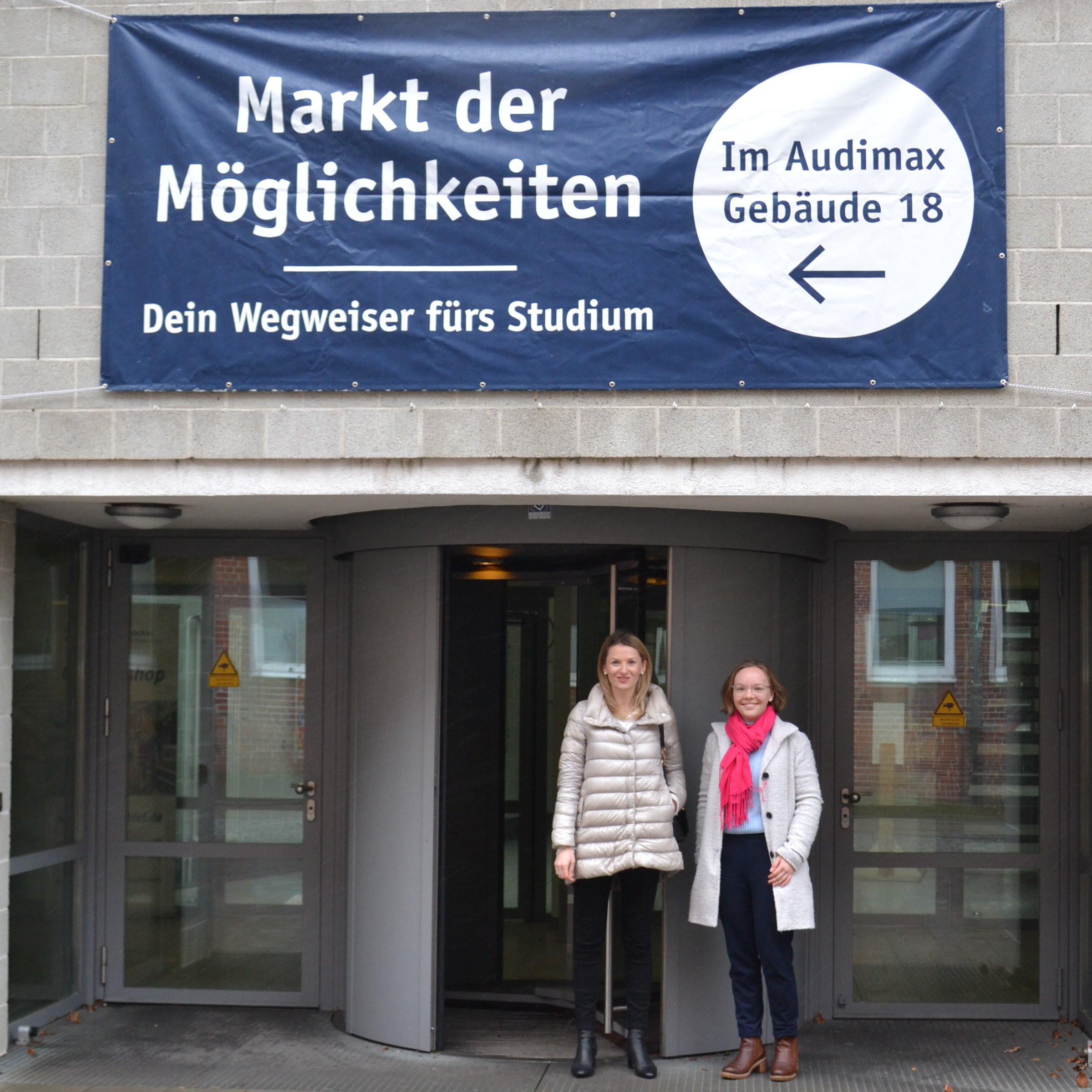 zwei Frauen vor einem Gebäude, hinter ihnen hängt ein Banner mit der Aufschrift: Markt der Möglichkeiten - Dein Wegweiser fürs Studium
