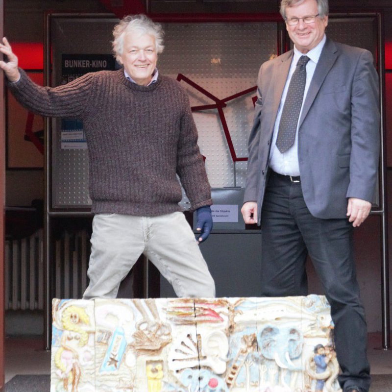 Zwei Männer, einer in Anzug, der ander in Strickpullover und Cordhose stehen auf einer Bühne und präsentieren Kunst.