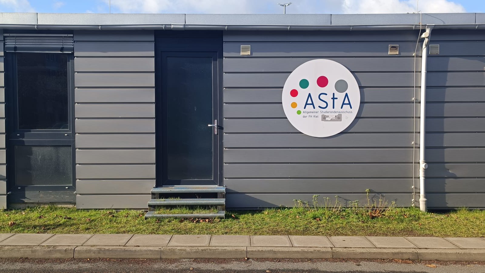 Ein graues Gebäude mit Holzverkleidung. Auf einem Schild steht man das Logo des AStA.