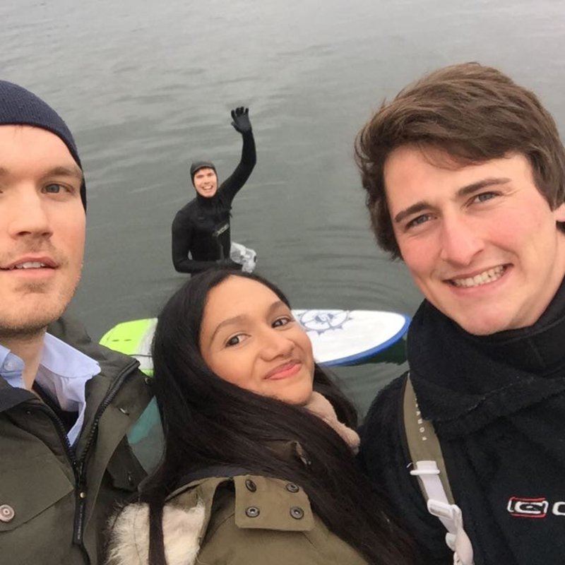 Zwei Männer und eine Frau machen ein Selfie am Ufer, auf dem Wasser winkt ein Mann in die Kamera.