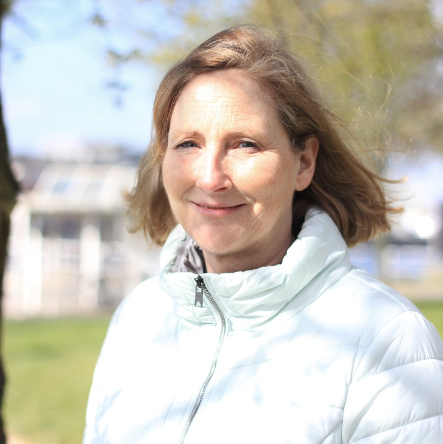 Eine Frau lächelt im Freien in die Kamera. Am rechten Bildrand ist ein Baumstamm zu erkennen, im Hintergrund ein Gebäude.