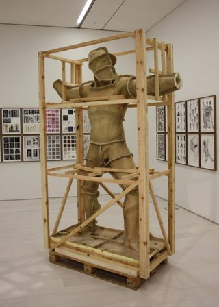 In einer Galerie steht eine Skulpur in Form eines Kriegeers, welche von einem Holzgerüst umgeben ist.