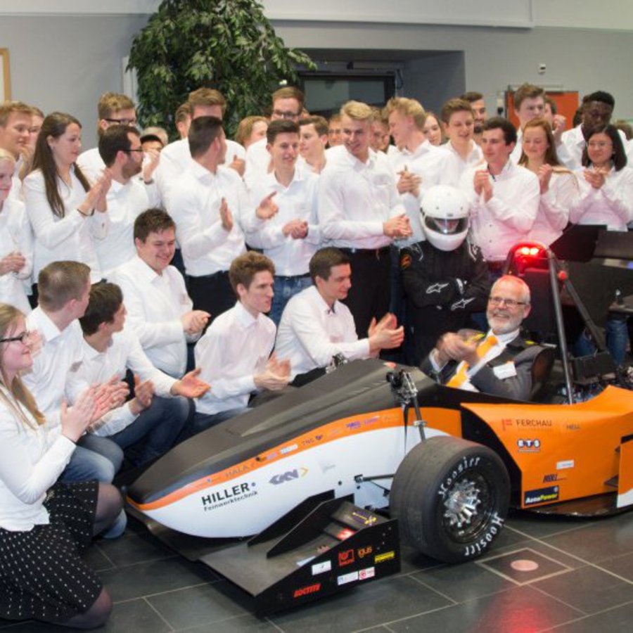 Ein älterer Mann in Anzug sitzt in einem Rennwagen. Er wird von einem applaudierenden Team in weißen Hemden umringt.