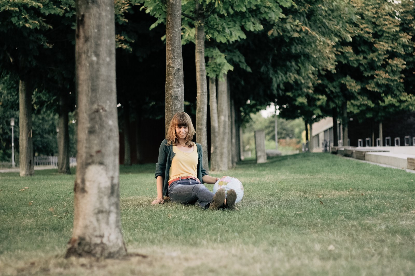 Eine Frau sitzt auf einem gepflegten Rasen und leht sich an einen dünnen Baum an. Neben ihr stützt ihre Hand auf einem aufblasbaren Globus.
