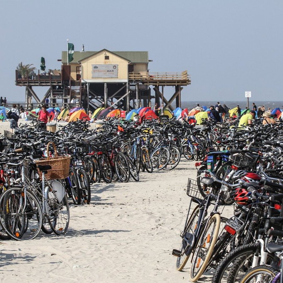 Der Strand von Sankt Peter Ording. Im Vordergrund stehen sehr viele Fahrräder, im Hintergrund sind ein Pfahlbau und die Kuppeln von kleinen Zelten zu sehen.