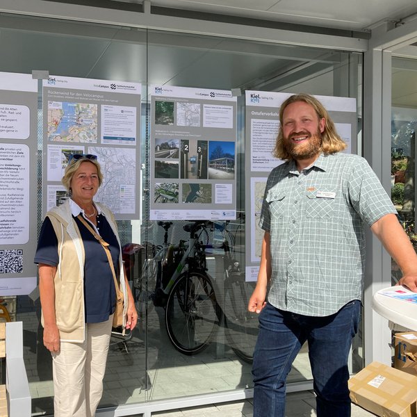 Eine Frau und ein Mann schauen lächelnd in die Kamera. Hinter ihnen sind Plakate an einer Glaswand aufgehängt