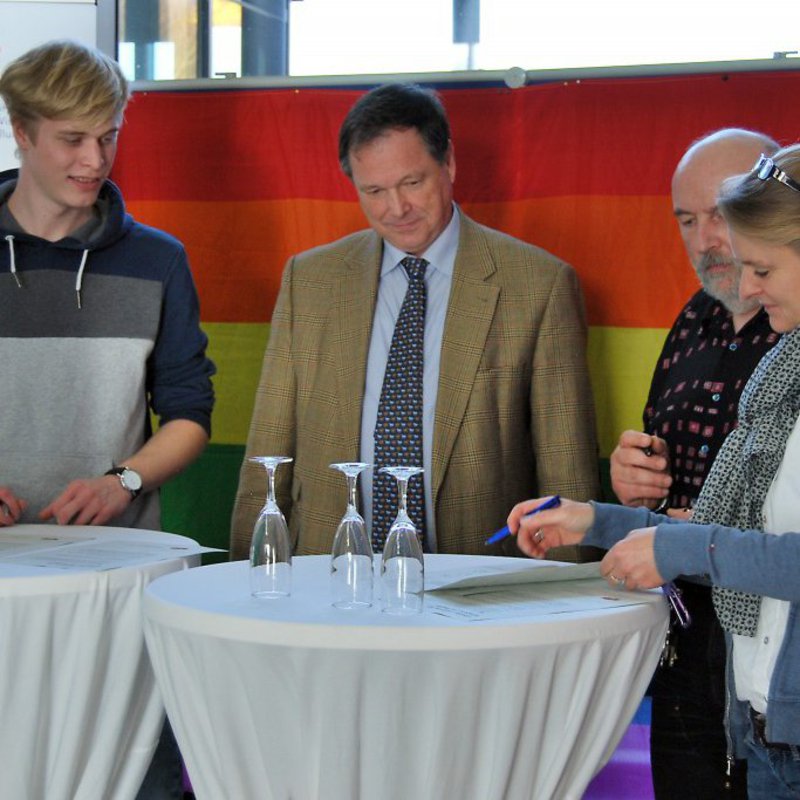 Zwei Männer und zwei Frauen stehen an einem Tisch und unterschreiben ein Schriftstück.