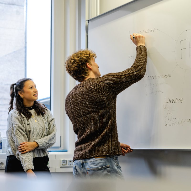 ein Mann beschreibt ein Whiteboard, zwei Frauen schauen ihm dabei über die Schulter