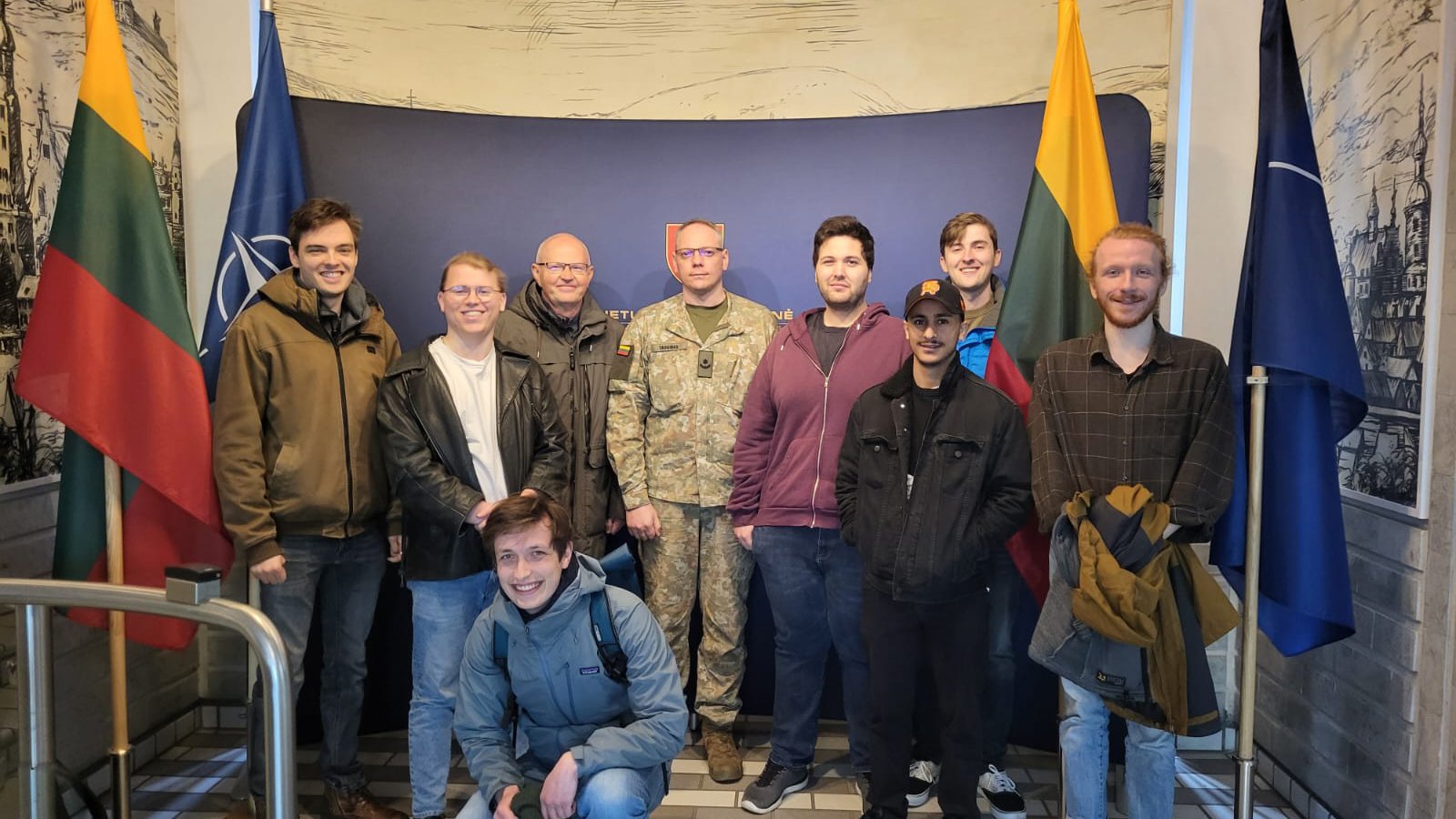 Acht Männer schauen lächelnd in die Kamera, einer von ihnen trägt eine Uniform in Tarnfarben, rechts und links der Gruppe sind litauische Flaggen zu sehen