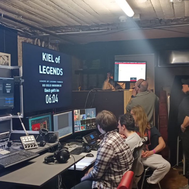 Auf einem großen Monitor sieht man den Titel des Turnieres "Kiel of Legends". Der Raum ist dunkel und es sind mehrere kleine Bildschirme und Tastaturen aufgebaut. Vor den Bildschirmen sitzen drei männlich gelesene Personen und im Hintergrund unterhalten sich drei Personen.