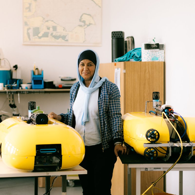 Professorin Sabah Badri-Höher steht zwischen zwei Tischen, auf denen die von ihr entwickelten Autonomen UNterwasserfahrzeuge liegen. 