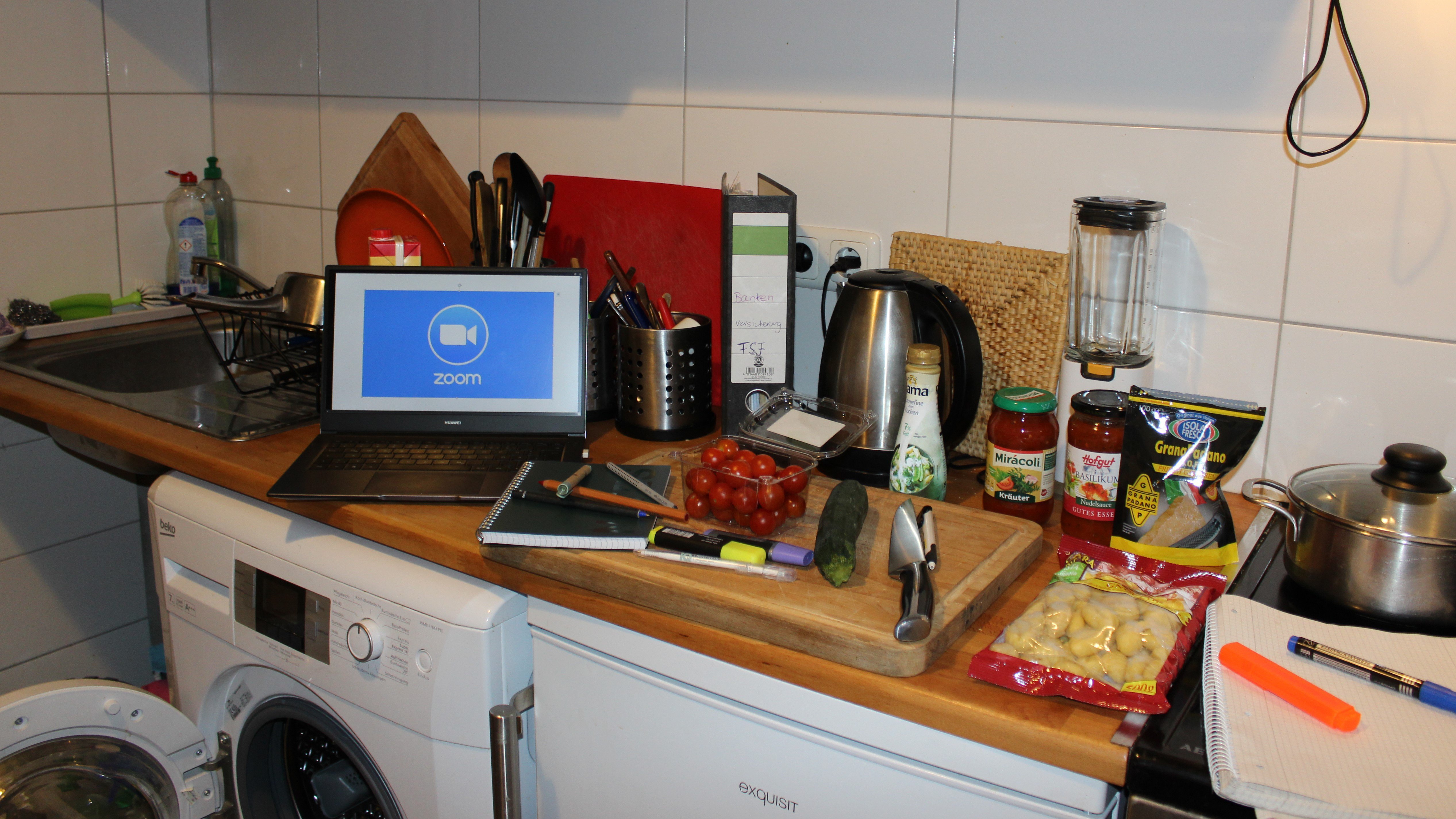 Laptop in der Küche auf der Arbeitsfläche, auf dem Display ist der Videokonferenz-Dienst Zoom geöffnet, auf und neben dem Schneidebrett liegen Lern- und Kochutensilien
