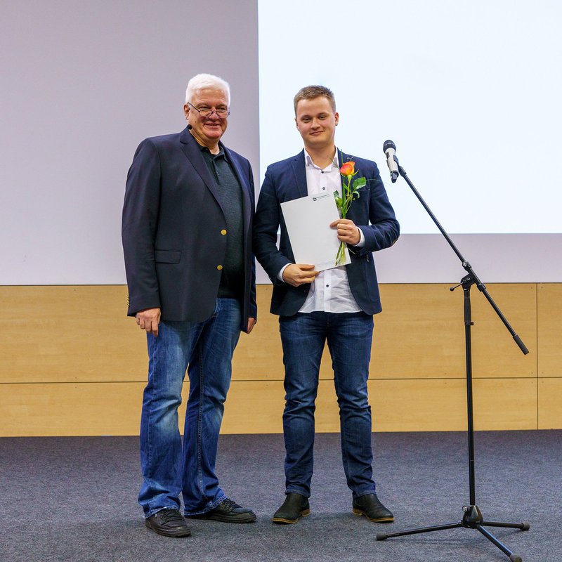 Auf einer Bühne stehen zwei Männer: ein älterer Mann links im Bild, Prof. Christian Hauck, und ein junger Mann, Silas Ketels, rechts im Bild. Beide lächeln in die Kamera, Silas Ketels hält eine Rose und seine Abschlussurkunde in der Hand. 
