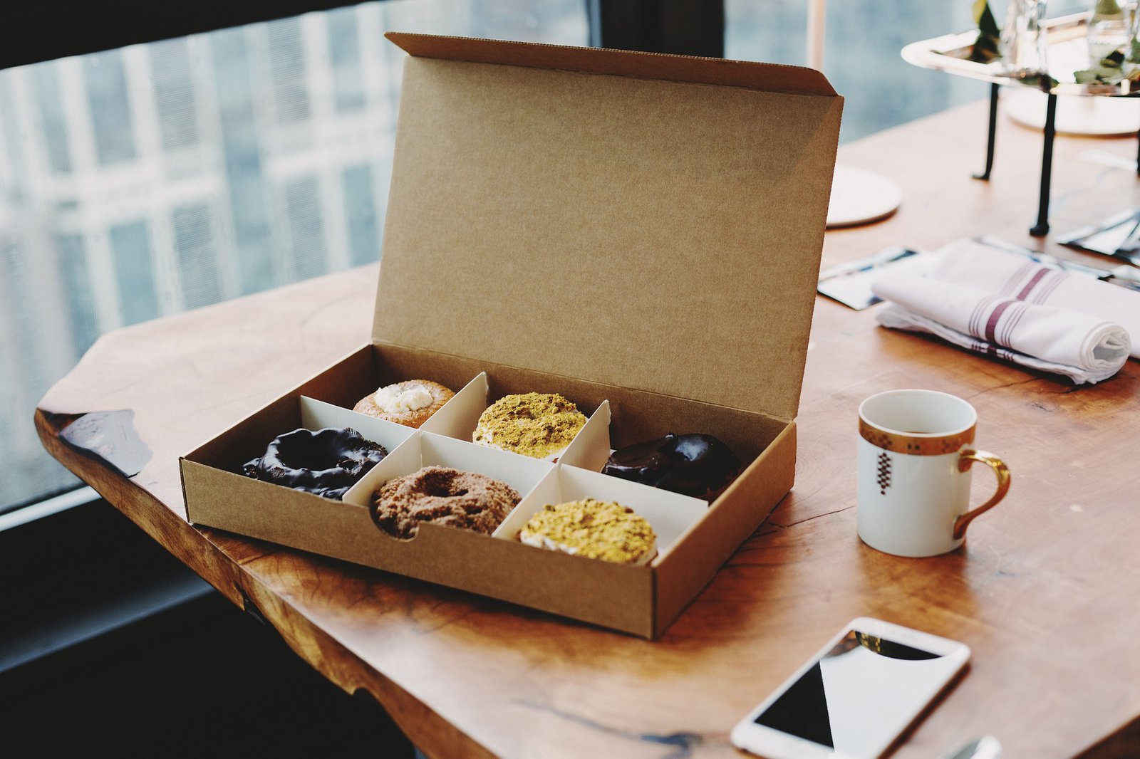 Eine Pappschachtel ist mit sechs Donuts befüllt und liegt auf einem Holztisch. Nebendran liegt ein Telefon und eine Kaffeetasse.