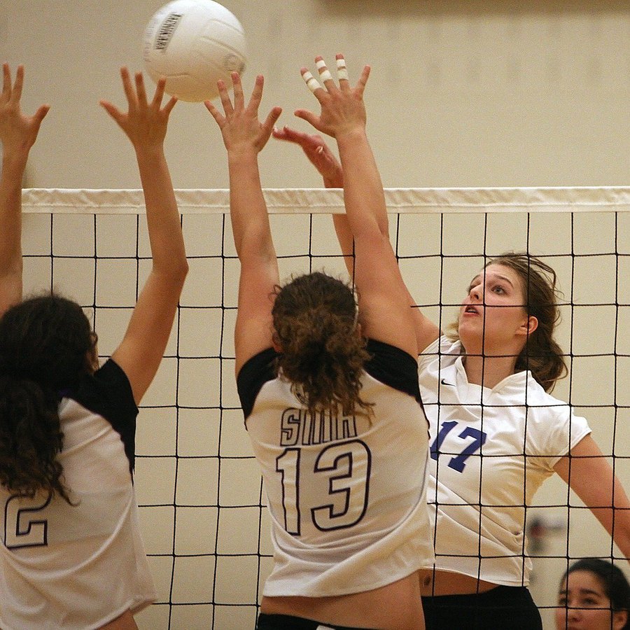 drei Volleyball spielende weiblich gelesene Personen, die sich um den Ball bemühen, der über ihren Köpfen schwebt. Es trennt sie ein Netz.