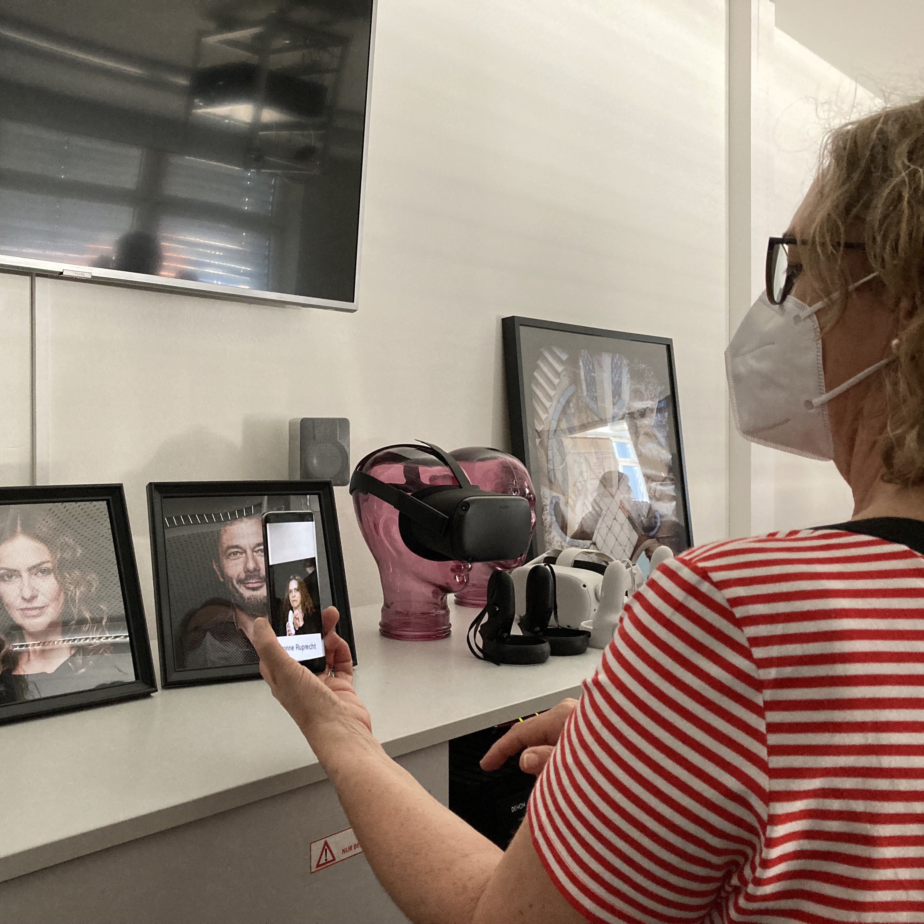Eine Frau schaut auf ein Smartphone, auf dem ein Foto ein Video einer Frau zu sehen ist. Im Hintergrund ist die Frau aus dem Video auf einem Foto zu sehen.