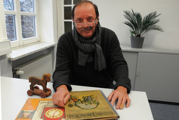 Ein Mann mit weisser Brille sitzt an einem Tisch, hält ein altes Buch in den Händen und lächelt in die Kamera.