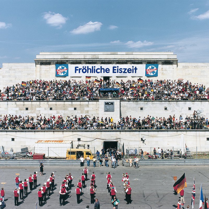 Das Bild zeigt einen Platz, auf dem sich eine Marschkapelle aufhält. Hinter dem Platz kommt ein zweistöckiges Plateau, auf dem sich Fams aufhalten und Deutschlandfahnen schwingen.