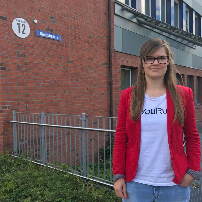 Eine junge Frau in rotem Blazer steht vor dem Gebäude des Fachbereichs Medien.