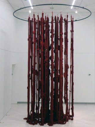 Eine von der Decke hängende Installation, bestehend aus einem oberhalb angebrachtem Ring und von ihm abgehenden roten dicken Stoffbändern.