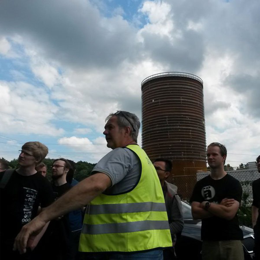 Ein Mann mit gelber Warnweste zeigt einer Gruppe Studierender einen Kessel.