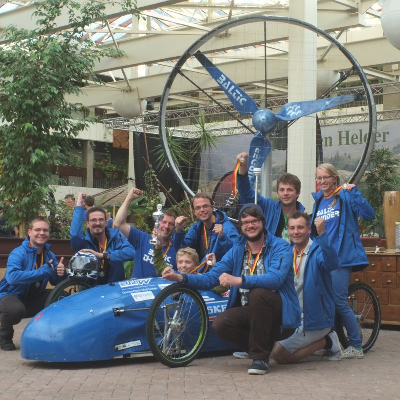 Ein Team in blauer Kleidung befindet sich jubelnd neben ihrem Gegenwindrennwagen.