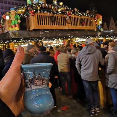 Eine Tasse mit Heißgetränk wird vor die Kamera gehalten, im Hintergrund ist ein Stand auf dem Weihnachtsmarkt zu sehen