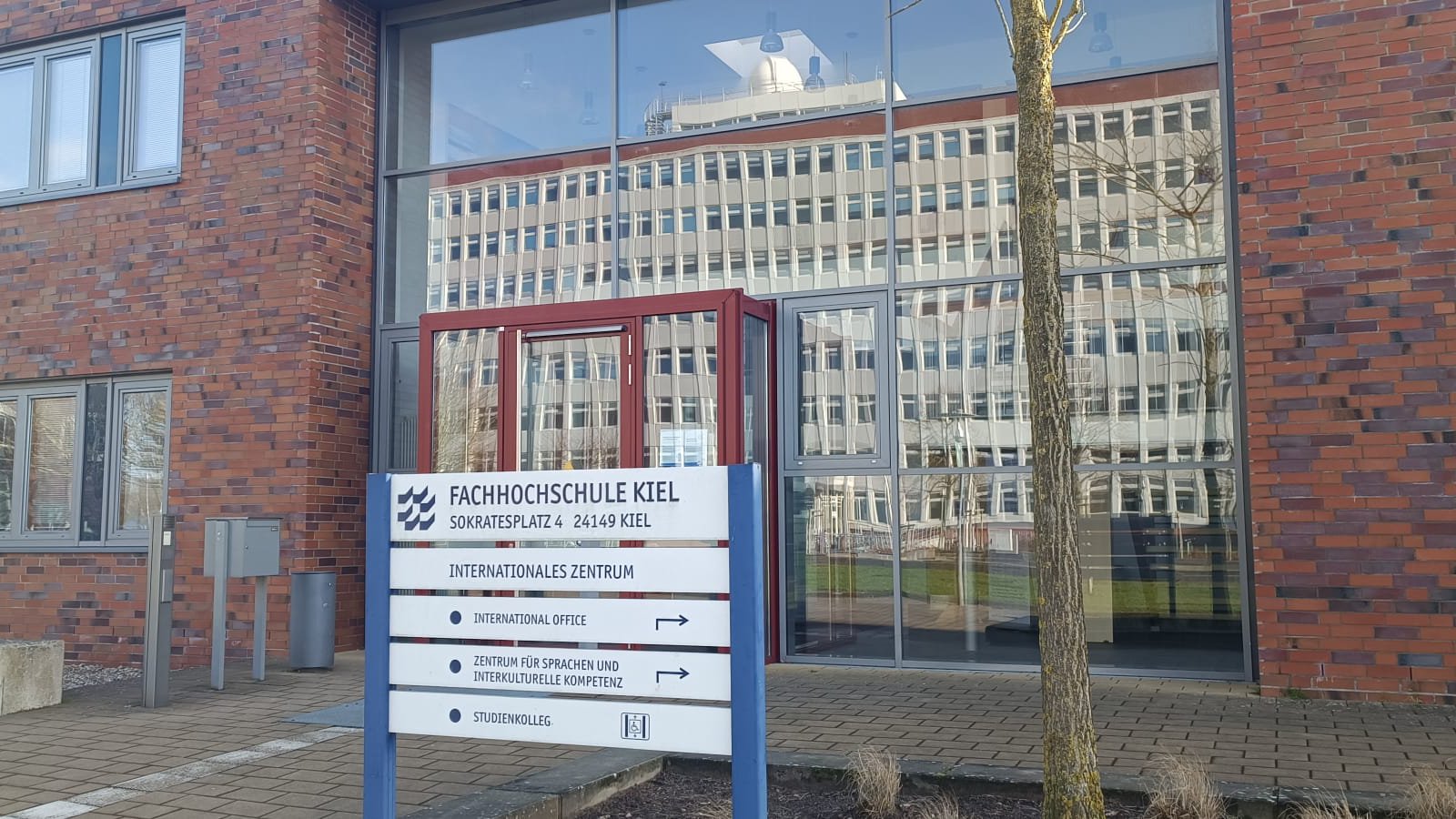 Ein rotbrauner Backstein-Bau mit großer Glasfassade, in welcher sich ein großes Gebäude spiegelt. Im Vordergrund steht ein Schild, welches das International Office ausweist.