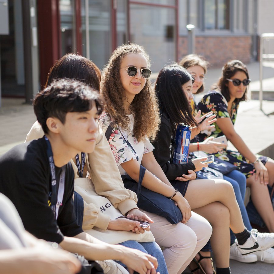 Eine Gruppe Student*innen sitzt auf den Stufen vor dem International Office in der Sonne.