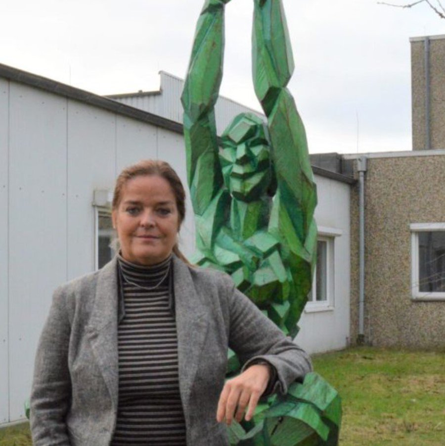 eine Frau im Rollkragenpullover und Blazer steht auf vor einer grünen Statue. Ihren Arm hält sie angelehnt an diese Statue. Im Hintergurnd sieht man den Teil eines Gebäudes.