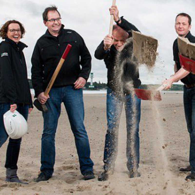 Eine Gruppe Menschen wirft Strandsand mit ihren Schaufeln hoch.