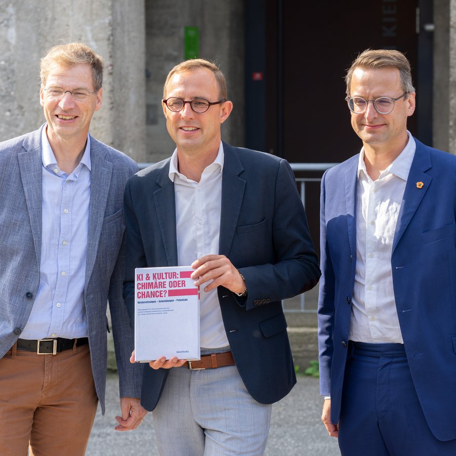 Drei Männer stehen vor dem Computermuseum der FH Kiel. Prof. Martin Lätzel (links) und Prof. Tobias Hochscherf (rechts). In der Mitte steht Digitalisierungsminister Dirk Schrödter und hält ein Buch in der Hand. 