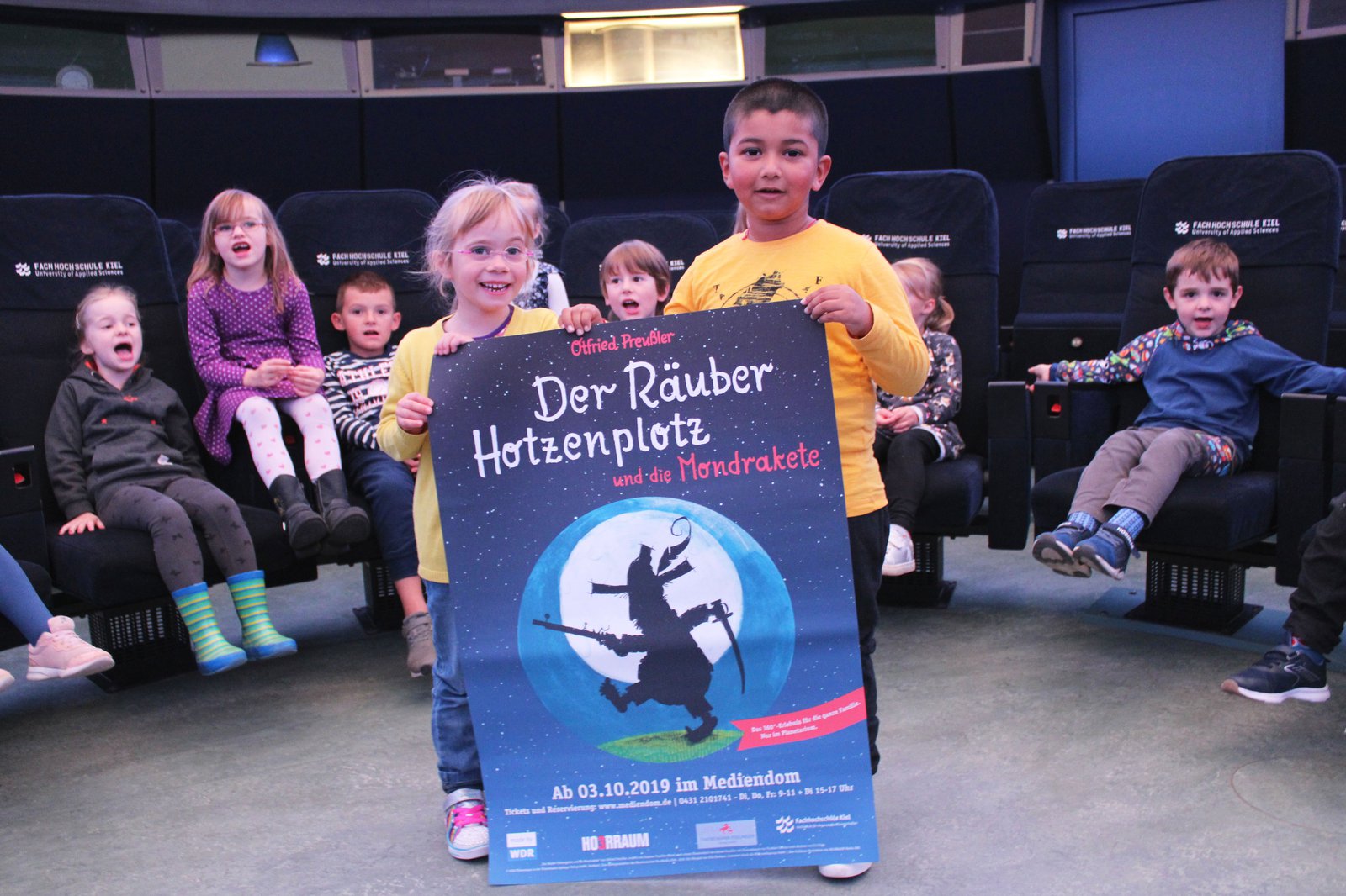 Zwei Kinder halten das Plakat zur Vorstellung "Räuber Hotzenplotz" in den Händen. Sie stehen in Mediendom und hinter ihnen befinden sich ihre Gruppenkameraden.