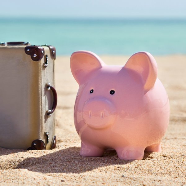 ein Sparschwein neben einem Alukoffer am Strand, dahinter Meer