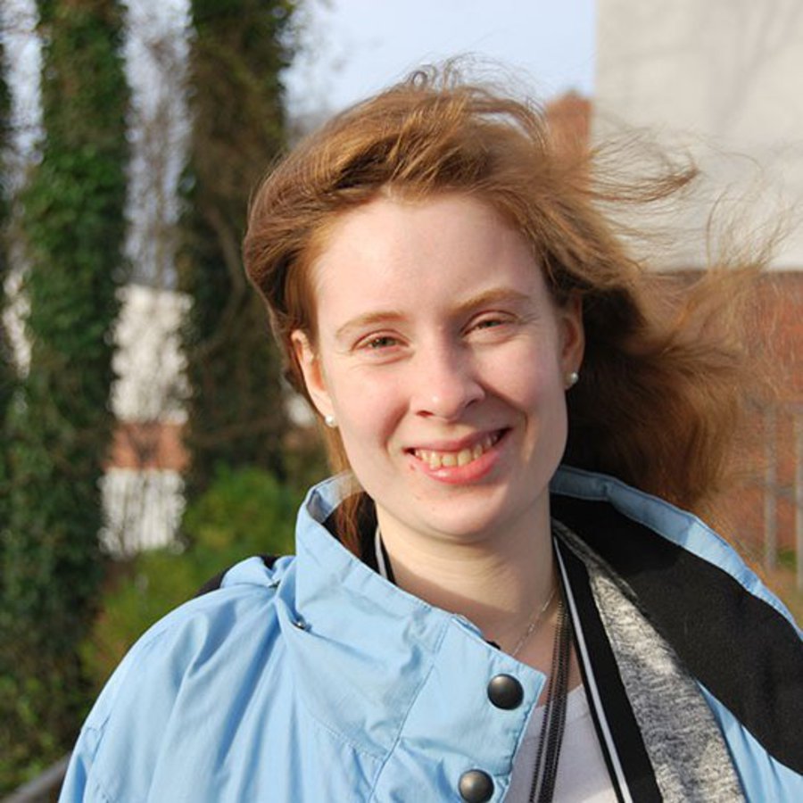 Ein Frau mit vom Wind verwehtem Haar, steht im Freien und lächelt freundlich in die Kamera.