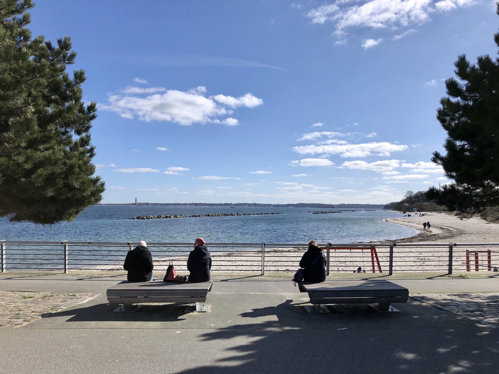 Das Bild zeigt Personen die auf Bänken sitzen und auf den vor ihnen liegenden Strand blicken.
