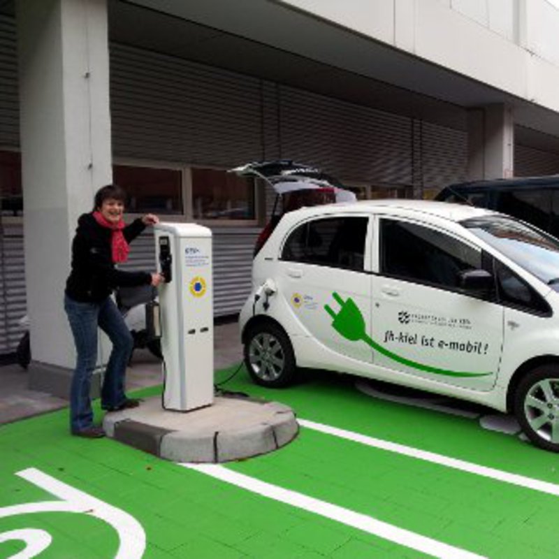 Eine Frau steht neben einem E-Auto, welches auf einem grünen Ladeparkplatz steht.