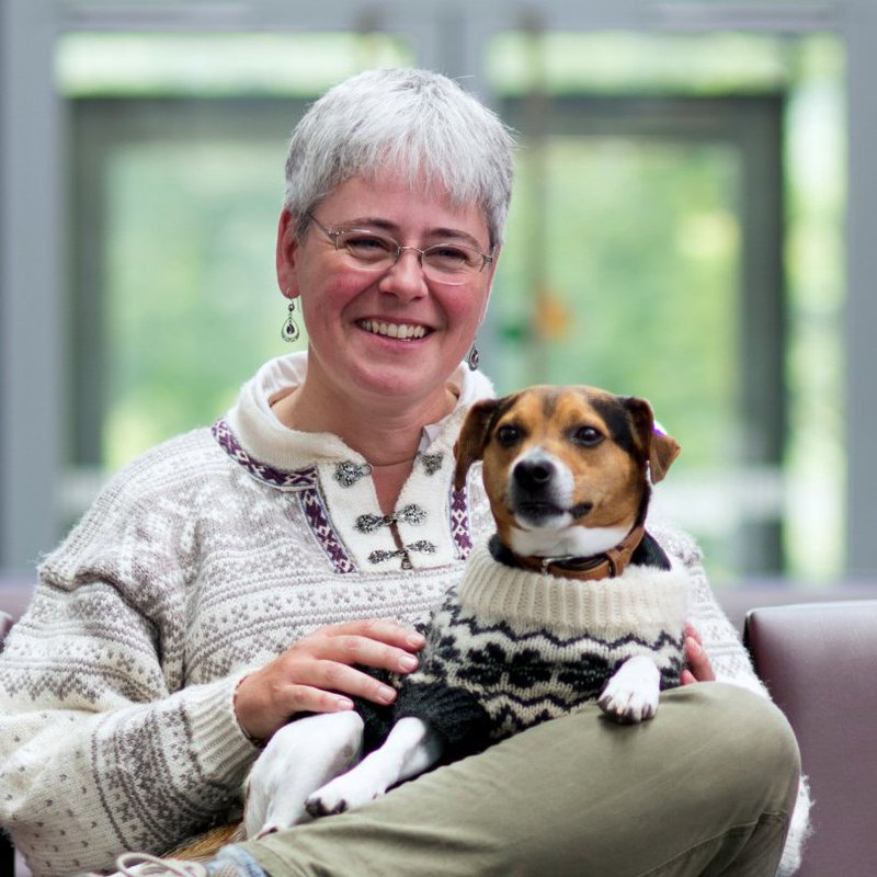 Eine ältere Frau hält ihren Hund auf dem Schoß und lächelt amüsiert in die Kamera.