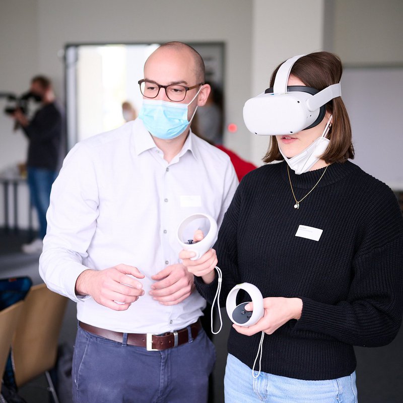 Eine Frau und ein Mann stehen in einem Raum, im Hintergrund sind weitere Personen zu sehen. Die Frau trägt eine Virtual-Reality-Brille und hält zwei Steuerungselemente in der Hand. Der Mann steht neben ihr und guckt in dieselbe Richtung. Foto: Andreas Diekötter.