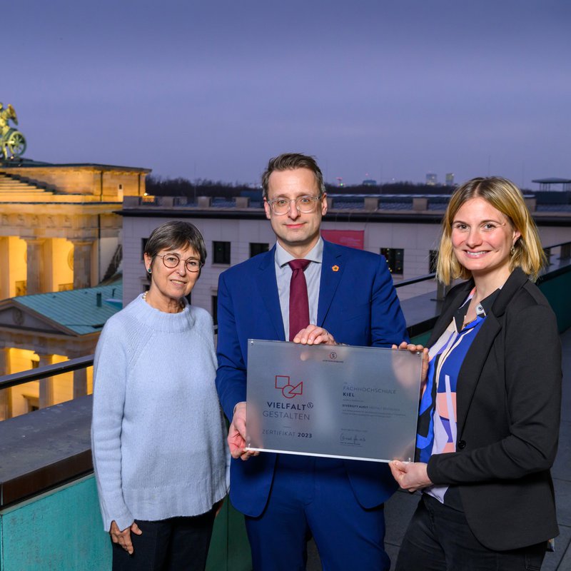 Prof. Katrin Hansen, Prof. Tobias Hochscherf und Alexa Magsaam stehen auf einem Balkon und halten eine Tafel mit der Aufschrift Vielfalt gestalten in Händen. Im Hintergrund ist das Brandenburger Tor zu sehen. 